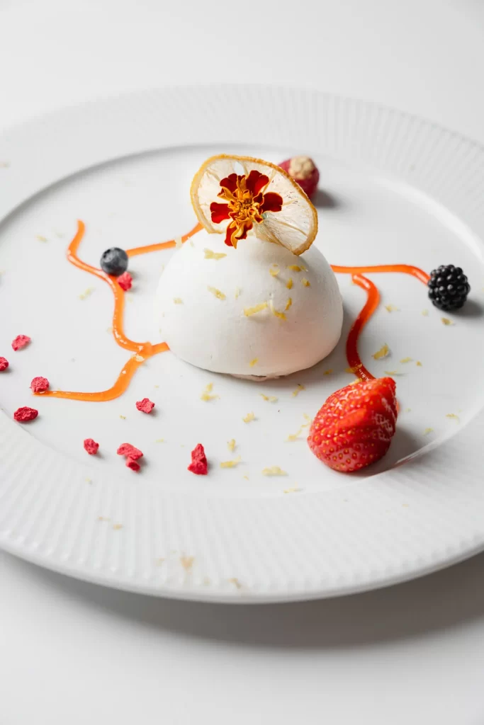 Dessert gourmet con mousse bianca decorata con frutta e un fiore essiccato su un piatto bianco.