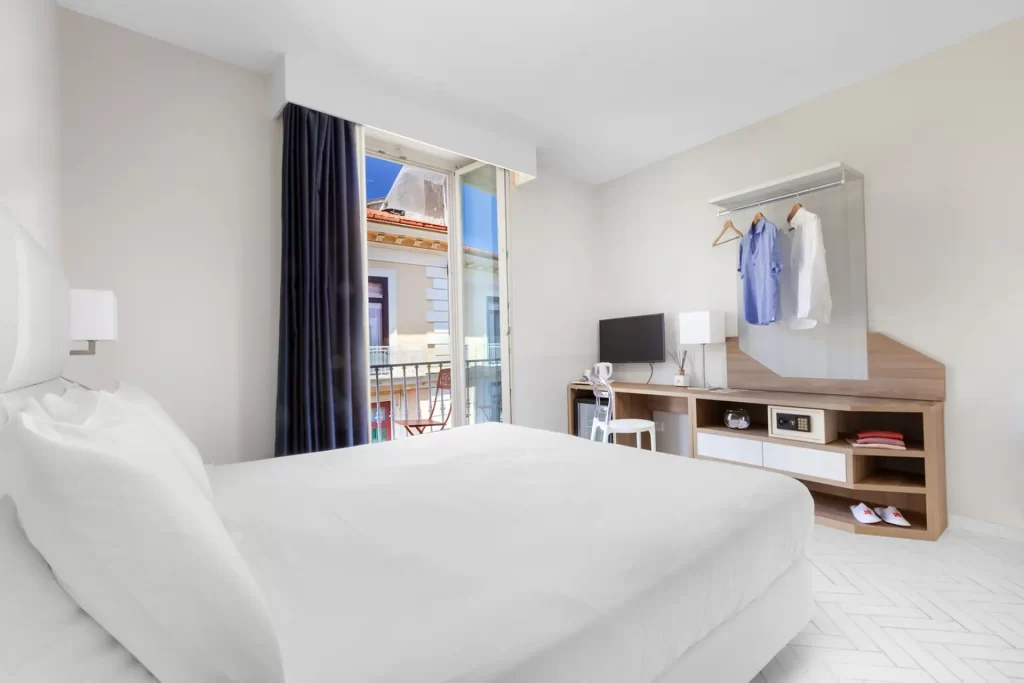 Camera moderna con letto bianco, scrivania e finestra aperta con vista sulla città.