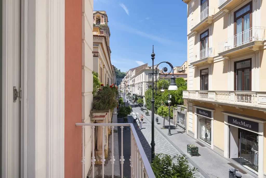 Street view from a balcony overlooking a sunny, tree-lined avenue with elegant buildings.