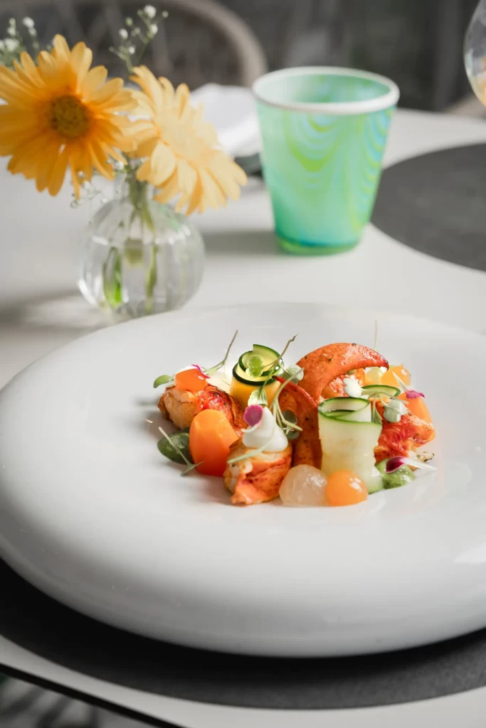 Un piatto elegante di aragosta con verdure su un tavolo decorato con fiori e un bicchiere colorato.