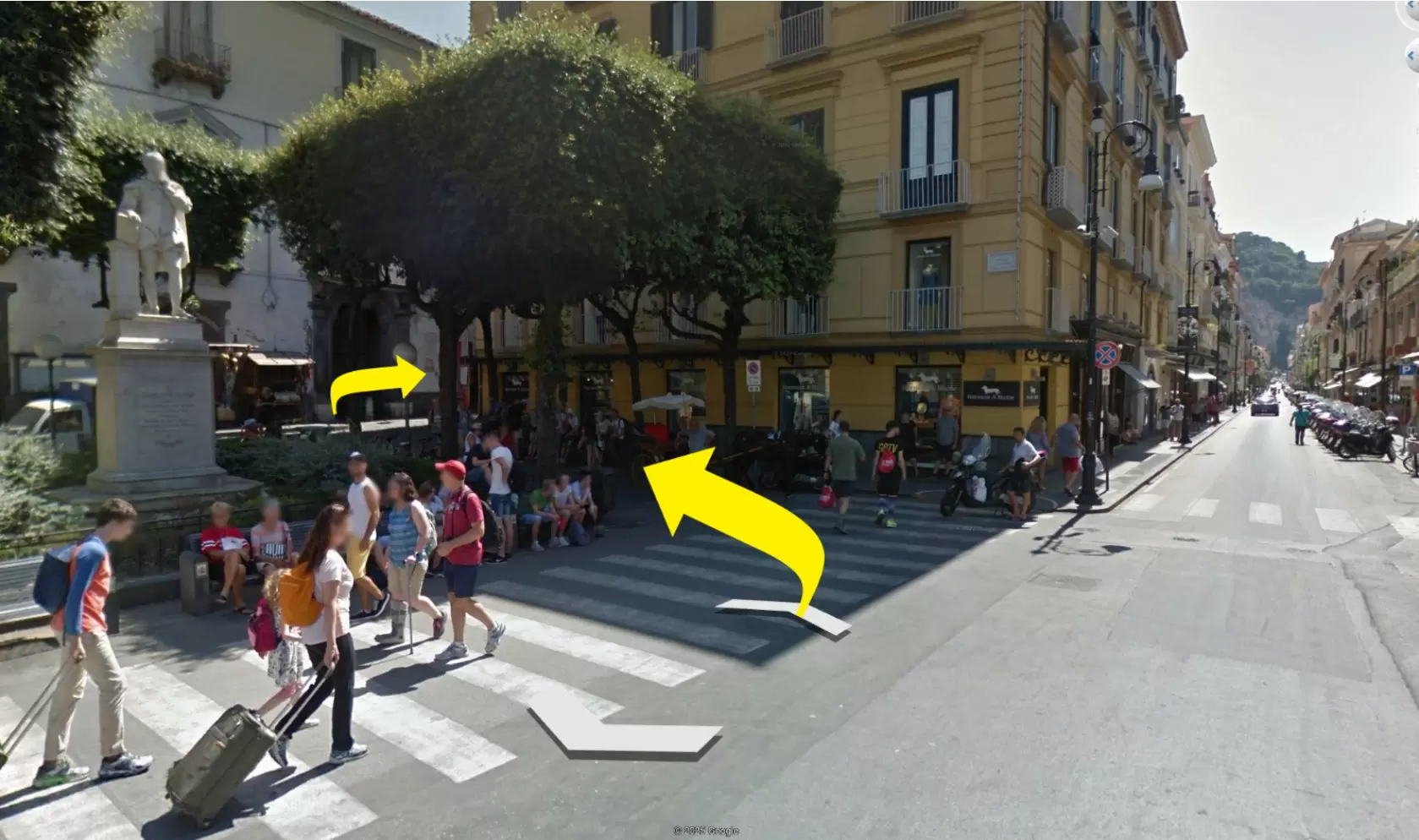 A busy street corner with people crossing a zebra crossing near a statue and a yellow building.