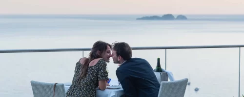 Coppia che cena romanticamente su una terrazza affacciata sul mare al tramonto.