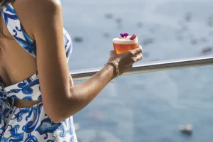 A woman in a blue patterned dress holds a drink on a balcony overlooking the ocean.