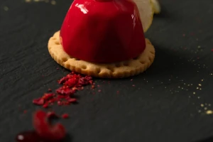 A red glazed dessert with a raspberry and gold leaf on a biscuit, served on a slate.