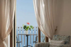 A cozy room with sheer curtains open to a balcony view of the sea and a potted plant on the table.