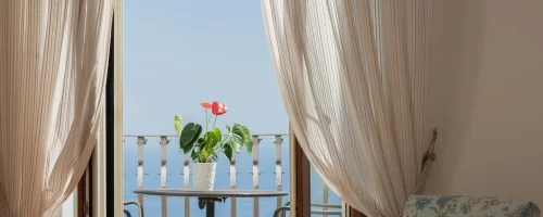 Interno luminoso con tende leggere, un balcone con vista sul mare e una pianta su un tavolino.