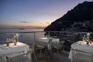 A seaside restaurant terrace at sunset with a view of the coast and illuminated hillside.