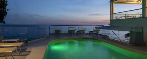 Terrazza con piscina illuminata e vista sul mare al tramonto.