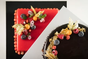 Two elegantly decorated cakes, one red and one chocolate, topped with fresh berries and golden leaves.