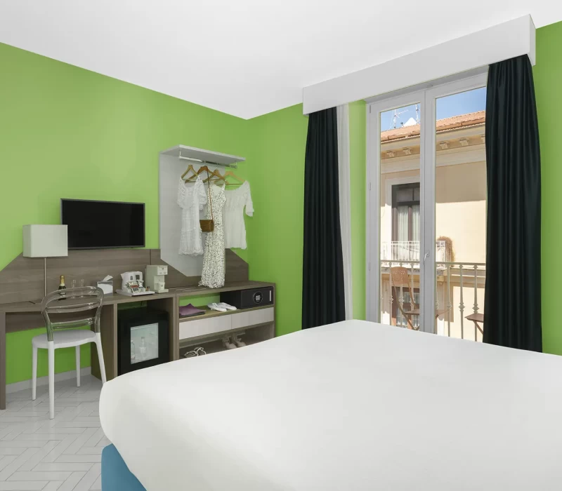 Camera d'albergo moderna con pareti verdi, letto bianco e una finestra che si affaccia su un balcone.