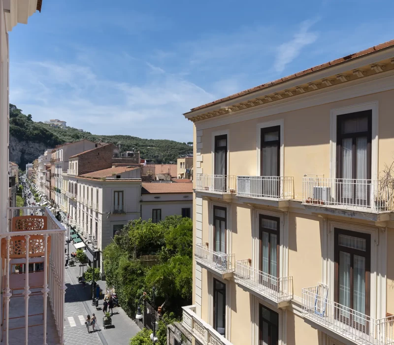 Vista di una strada alberata con edifici storici e montagne sullo sfondo in una giornata soleggiata.