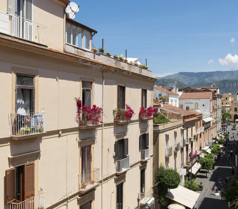 Strada italiana con edifici storici, balconi fioriti e vista sulle montagne.