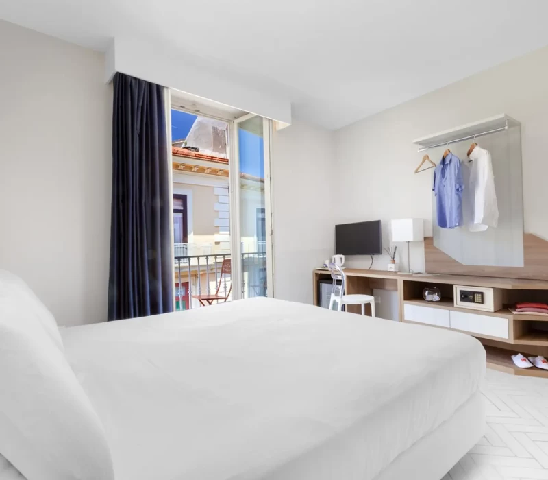 Camera moderna con letto bianco, scrivania e finestra aperta con vista sulla città.