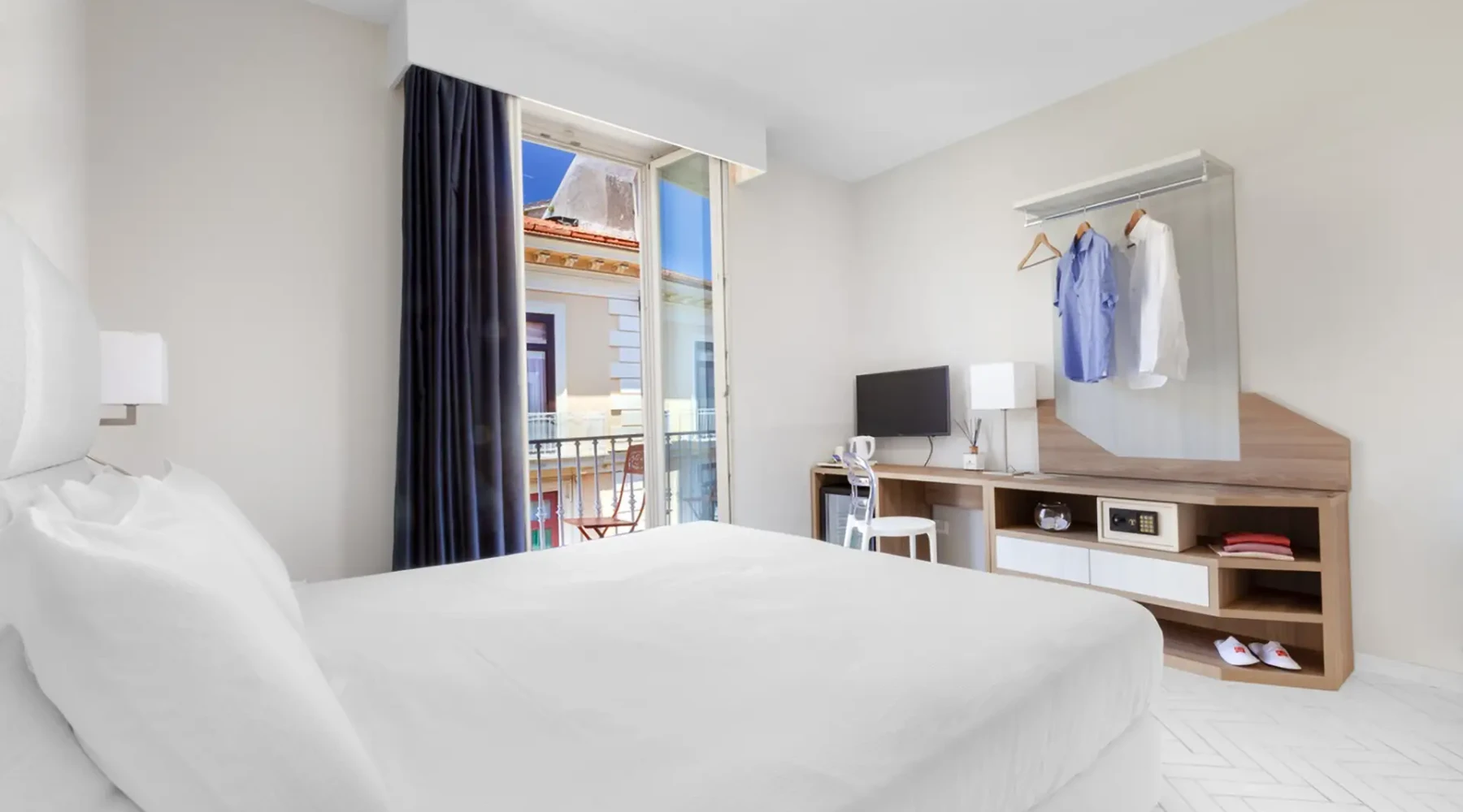 Bright hotel room with a bed, desk, wardrobe, and open balcony.