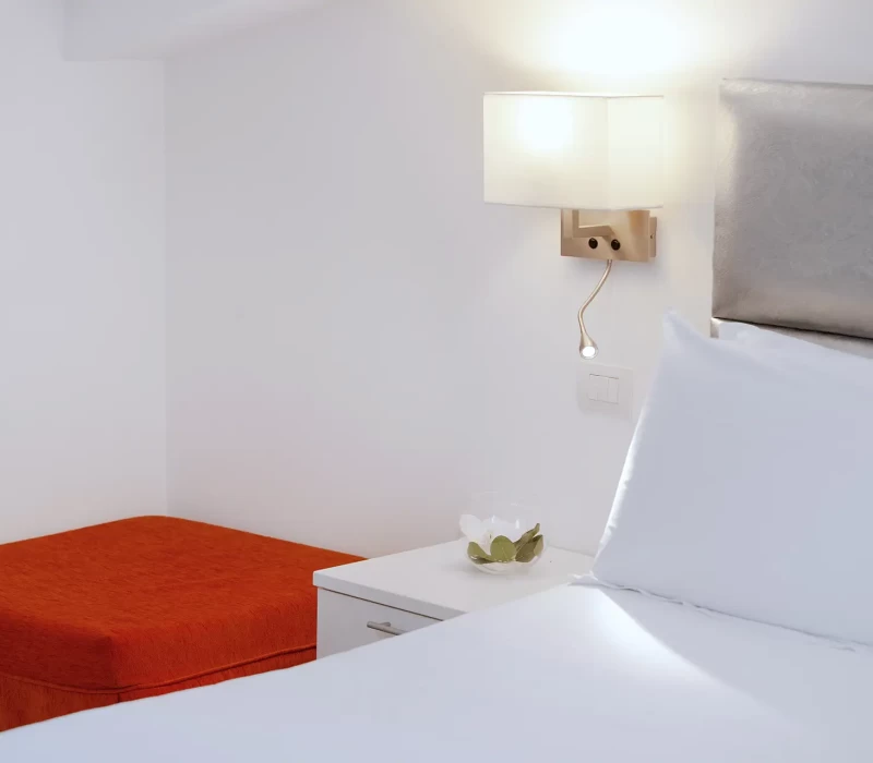 Camera da letto minimalista con lampada da parete, testiera imbottita, comodino bianco e pouf arancione.