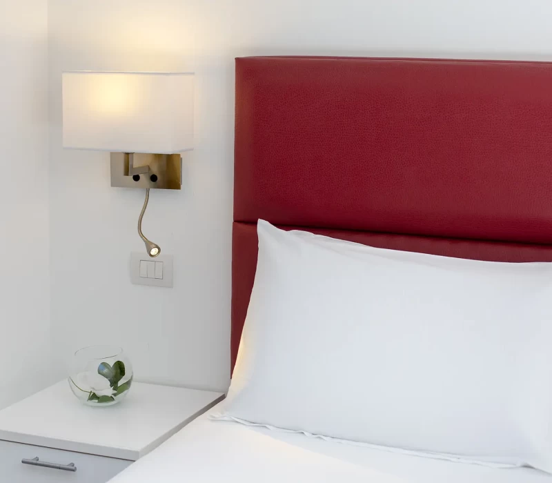 Letto con testiera rossa, lampada da parete e comodino bianco con vaso di fiori.