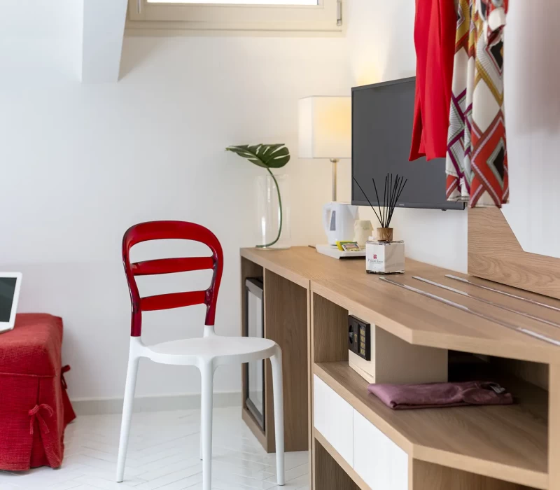 Angolo di una camera moderna con sedia rossa, laptop, TV e accessori su un mobile di legno.