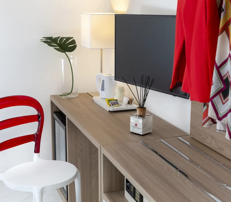 Angolo di una stanza moderna con sedia bianca e rossa, scrivania di legno, lampada, televisore e abbigliamento appeso.